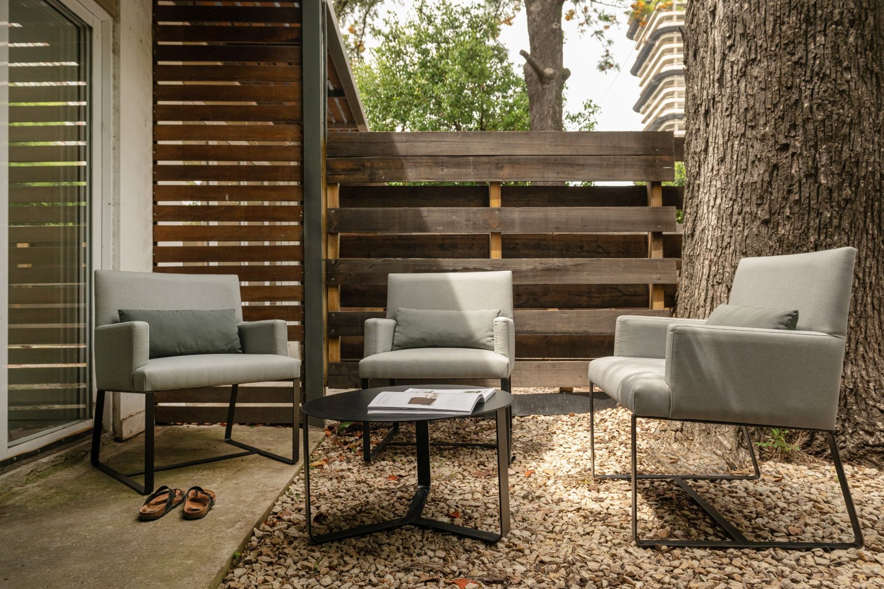 solsken – piezas que se despiden Tres sillones tapizados en gris claro con estructura de metal negro y mesa ratona redonda negra, en un patio con piso de piedra, cerco de madera y un árbol.